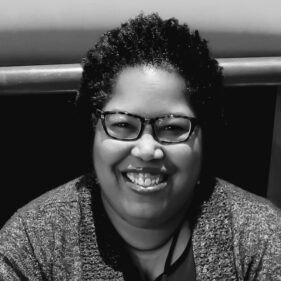 A middle-aged African American woman with short, dark hair, a grey sweater, and glasses smiles in front of a dark backdrop