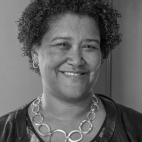 an older African-American woman with short black hair, a blue shirt, earrings, and a decorative necklace smiles in front of a wall painted grey and red.