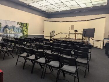 image of a large, beige-colored room with rows of black chairs, a small stage, and a TV monitor. 