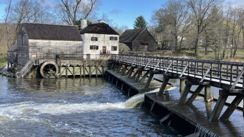 In the foreground is a long wooden footbridge crossing a river. In the background are three buildings: a historic mill and waterwheel made of wood, a white-washed three story colonial manor house, and a wooden barn